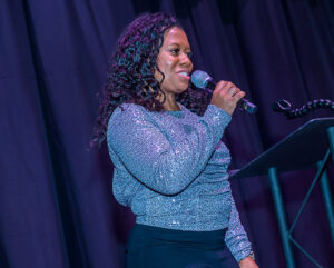 A woman in a sparkly top speaks into a handheld microphone on a stage with a dark curtain behind her.