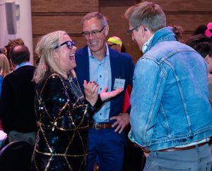 Three people share a laugh at a social event, one woman in a sparkly outfit and two men in casual and business attire.