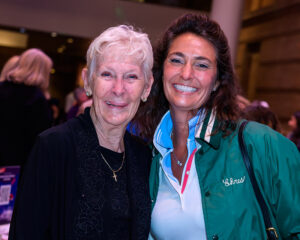 Two women smiling, one older in black, the other younger in a green jacket, standing close in an indoor setting.