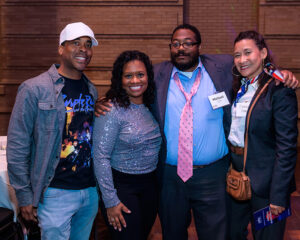 Four people posing for a group photo at an indoor event, smiling and standing close together.