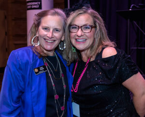 Two smiling women at an event, one wearing a name badge and both dressed up, one in a black outfit with accessories.