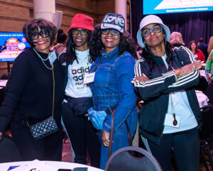 Four women, wearing casual and sporty attire, pose together at an indoor event with a bright screen in the background.