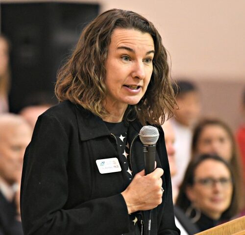 Woman speaking into a microphone at a podium with people seated in the background.