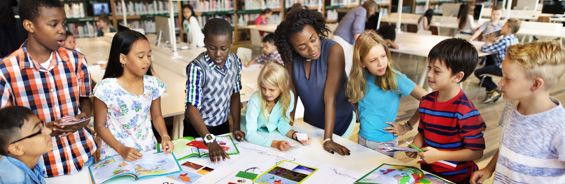 A teacher and diverse group of students interact around a large table with maps and books in a library setting.