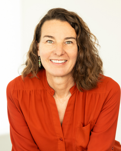 Smiling person with wavy brown hair, wearing a red blouse and earrings, against a light background.