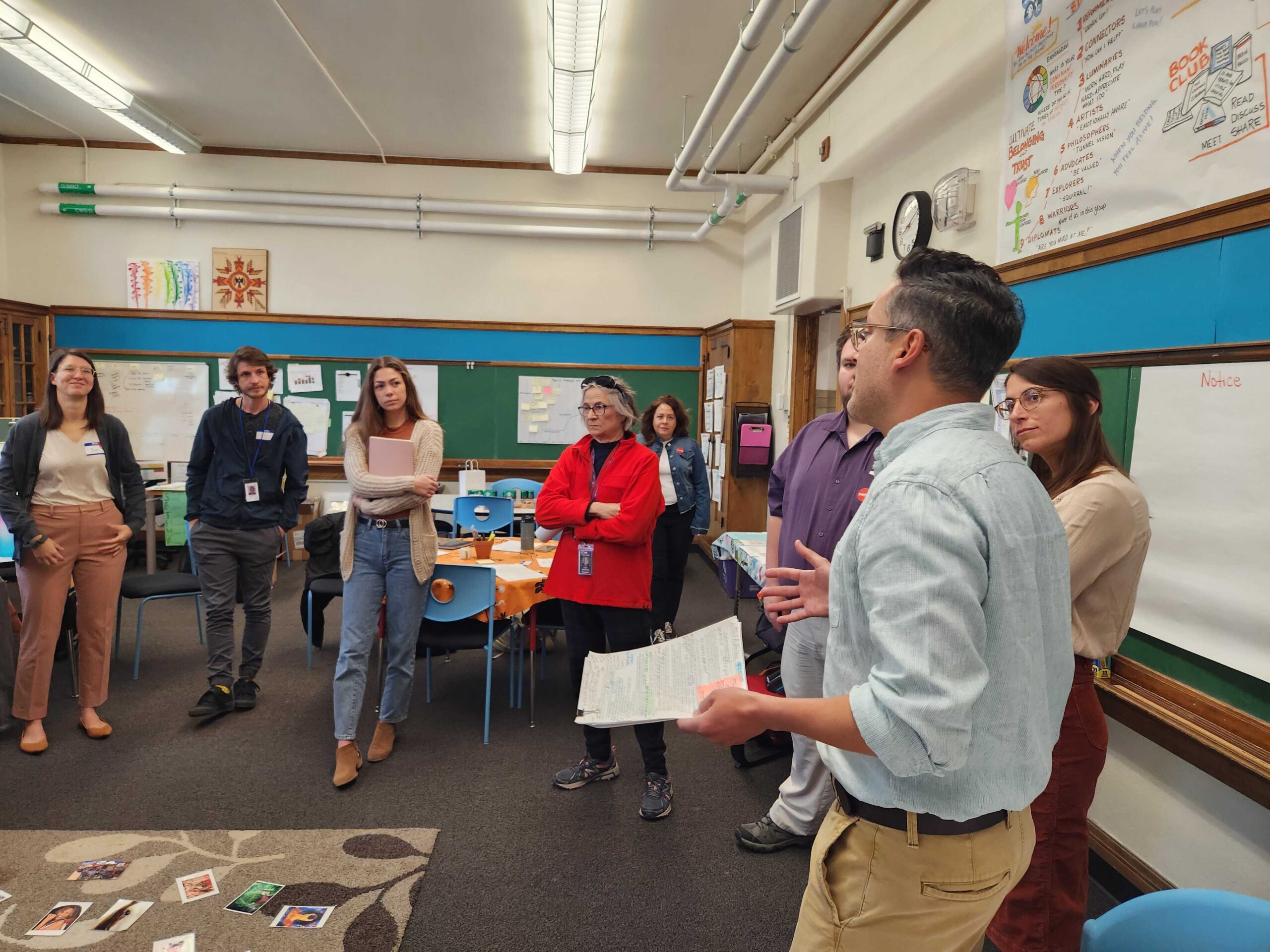 A group of people stand and listen to a speaker in a classroom, with posters and notes on the walls.
