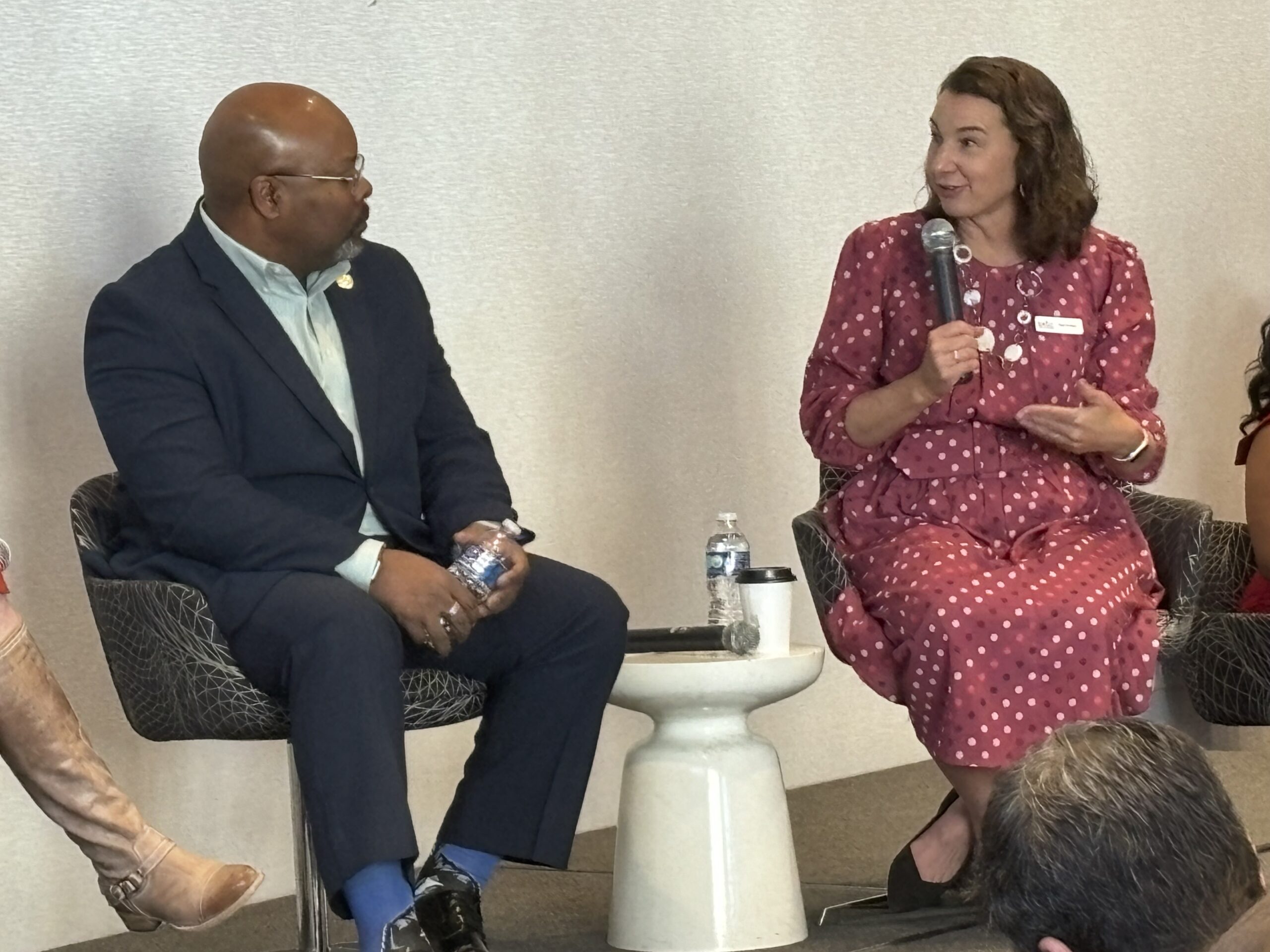 Two people sit on chairs, engaged in a discussion; one is holding a microphone and speaking.