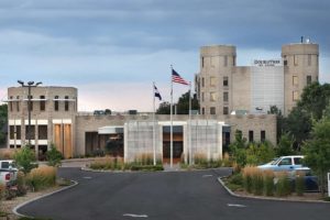 A modern hospital with two flagpoles in front, surrounded by parking spaces, under a cloudy sky.