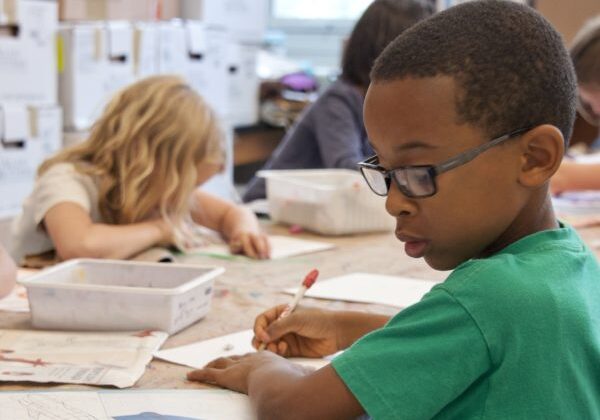 Children are seated at a classroom table, focused on writing and drawing activities.