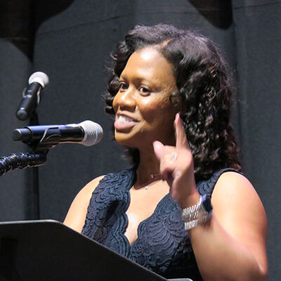 A woman speaks passionately at a podium, with a microphone in front of her and her finger pointed upward.