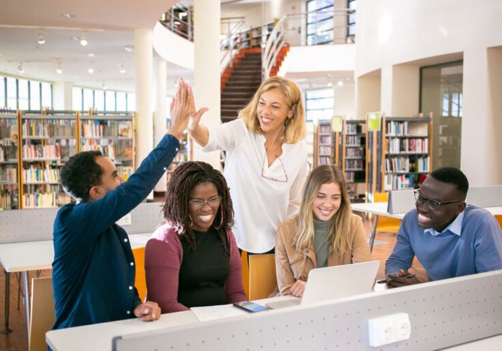 A diverse group of people high-fiving and smiling while working together in a library with laptops on the table.