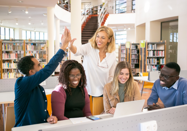 Group of five people in a library, smiling and high-fiving around a laptop, with bookshelves in the background.