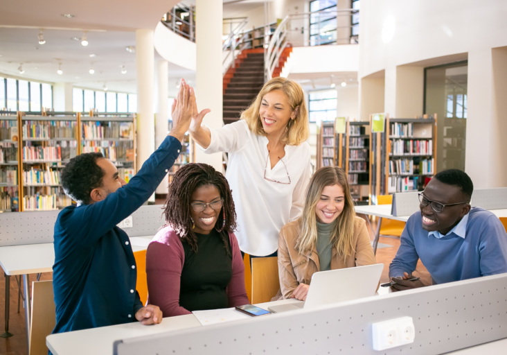 Group of five people in a library, smiling and high-fiving around a laptop, with bookshelves in the background.