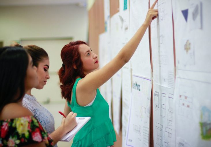 Three women stand and examine papers on a bulletin board; one points while another takes notes.