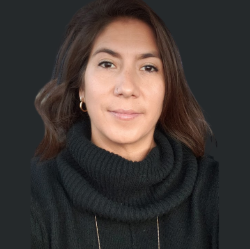 A person with long brown hair, wearing a black turtleneck sweater, poses against a plain dark background.